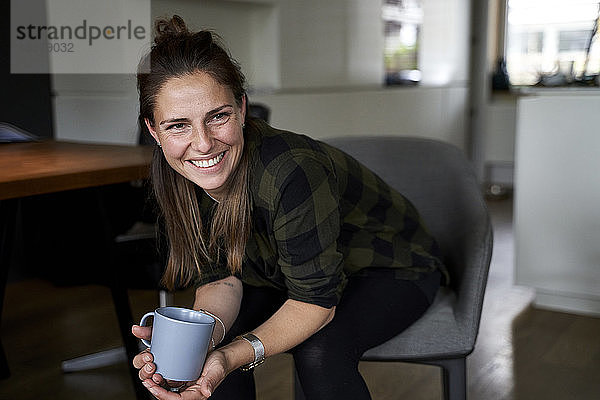 Lächelnde Frau  die eine Kaffeetasse hält und wegschaut  während sie auf einem Stuhl zu Hause sitzt