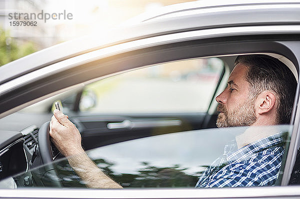 Seitenansicht eines gut aussehenden Mannes  der im Auto sitzend ein Mobiltelefon benutzt