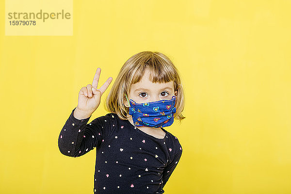 Porträt eines niedlichen blonden Mädchens  das eine Gesichtsmaske trägt und ein Friedenszeichen vor gelbem Hintergrund gestikuliert