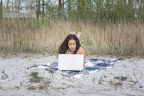 Junge Frau liegt auf einer Decke am Strand und benutzt einen Laptop