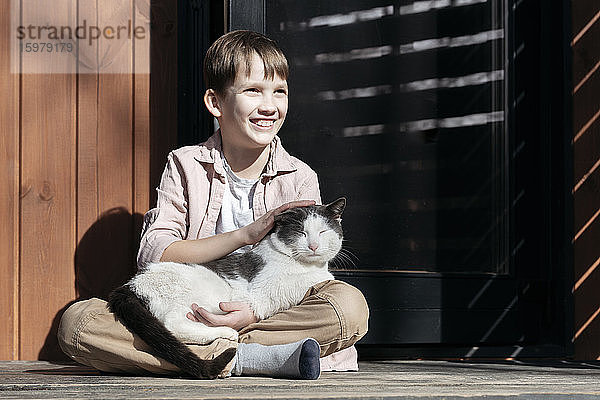 Lächelnder Junge  der wegschaut  während er eine Katze streichelt  die sich auf seiner Veranda an einem sonnigen Tag ausruht  Tarusa  Russland