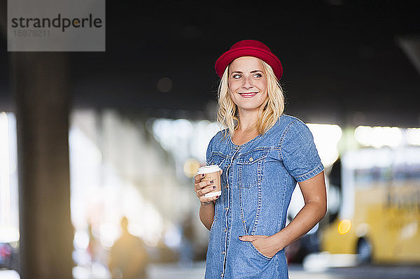 Porträt einer lächelnden blonden Frau mit Kaffee zum Mitnehmen in Jeanskleid und rotem Hut