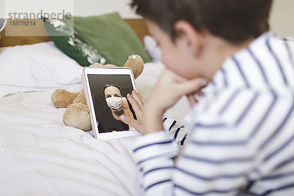 Junge auf dem Bett liegend  der mit seiner Mutter  die eine Schutzmaske trägt  einen Videogespräch auf einem digitalen Tablet führt