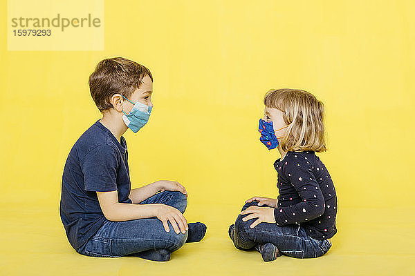 Ganzkörper-Seitenansicht von Geschwistern mit Gesichtsmasken  die vor einem gelben Hintergrund sitzen