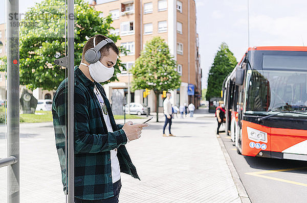 Junger Mann mit Kopfhörern und Schutzmaske  der sein Smartphone benutzt  während er an einer Bushaltestelle wartet  Spanien