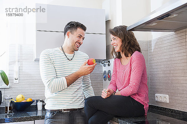 Glücklicher Mann  der einen Apfel in der Hand hält und eine Frau betrachtet  die auf dem Küchentisch sitzt