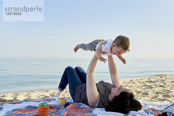 Mutter hebt glücklichen Sohn auf  während er auf einer Decke am Strand liegt