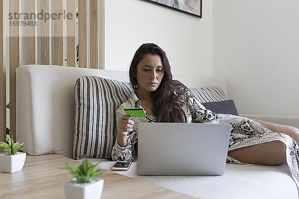 Schöne junge Frau hält Kreditkarte für Online-Shopping durch Laptop  während auf dem Sofa zu Hause liegen