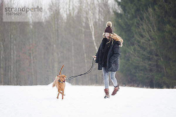 Glückliche junge Frau läuft mit Hund auf Schnee gegen Bäume im Wald im Winter