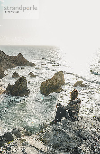Junge Frau sitzt auf einer Felsformation und schaut auf das Meer  Praia da Ursa  Lisboa  Portugal