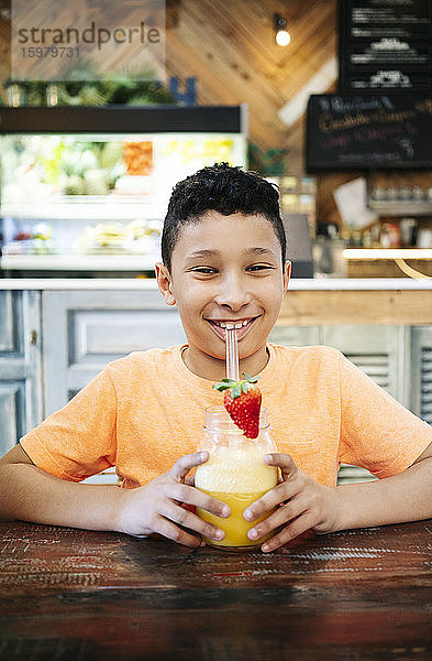 Porträt eines lächelnden Jungen  der einen frischen Smoothie in der Hand hält  während er am Tisch im Restaurant steht