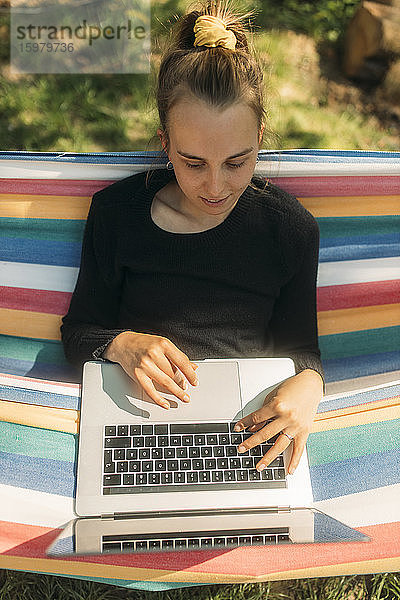 Junge Frau  die einen Laptop benutzt  während sie in einer Hängematte im Garten sitzt  während eines sonnigen Tages