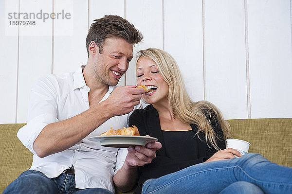 Lächelnder Mann füttert seine Freundin mit einem Croissant  während er zu Hause auf dem Sofa sitzt