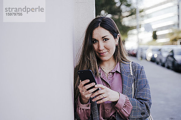 Porträt einer lächelnden Frau mit Smartphone  die an der Wand lehnt