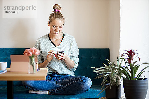 Lächelnde blonde Frau sitzt auf einer Bank zu Hause und schaut auf ihr Handy