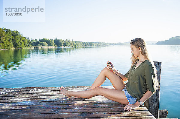 Junge Frau sitzt auf einem Steg am See und schaut auf ihr Handy