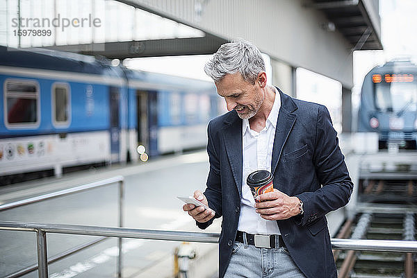 Geschäftsmann mit Kaffee in der Hand und Smartphone im Bahnhofsbereich