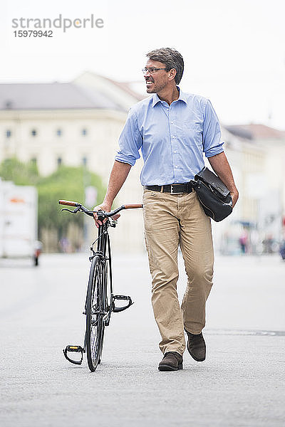 Lächelnder Geschäftsmann schaut weg  während er mit dem Fahrrad auf der Straße in der Stadt spazieren geht
