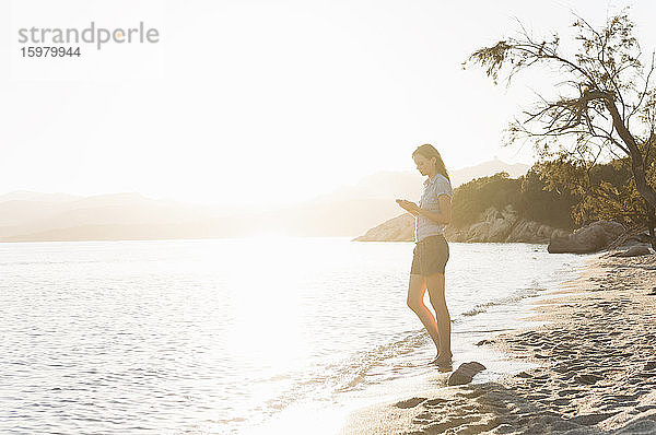 Frau steht bei Sonnenuntergang am Meer und schaut auf ihr Handy  Sardinien  Italien