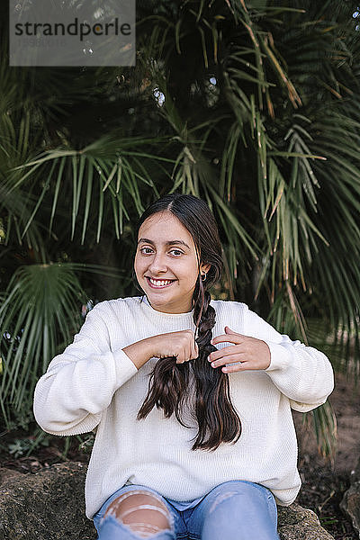 Porträt eines lächelnden Teenager-Mädchens  das sein langes braunes Haar flechtet  während es an einer Palme im Park sitzt