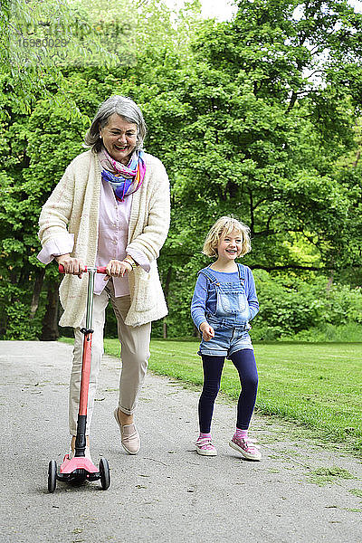 Aktive ältere Frau fährt mit dem Roller  während ihre Enkelin im Park spazieren geht