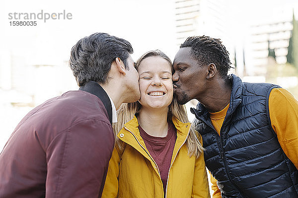 Zwei junge Männer küssen eine lächelnde junge Frau im Freien