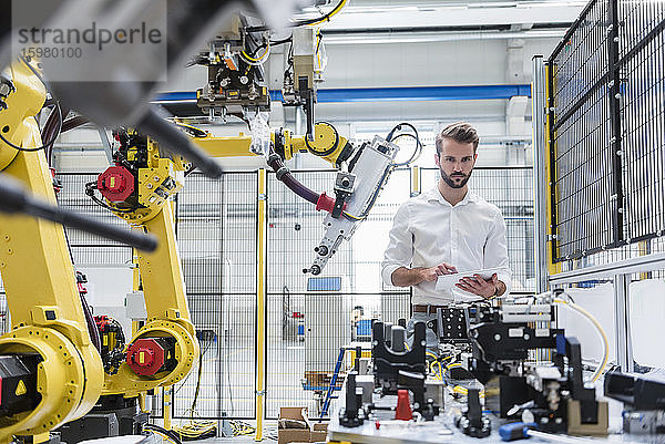 Junger Ingenieur hält ein digitales Tablet in der Hand und betrachtet Maschinen in einer Fabrik