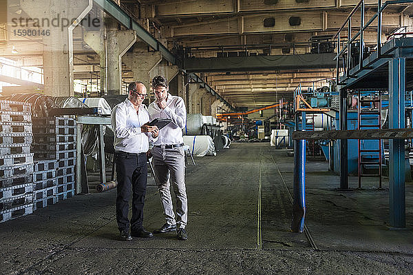 Zwei Geschäftsleute bei einer Besprechung mit Blick auf ein Tablet in einer Fabrikhalle
