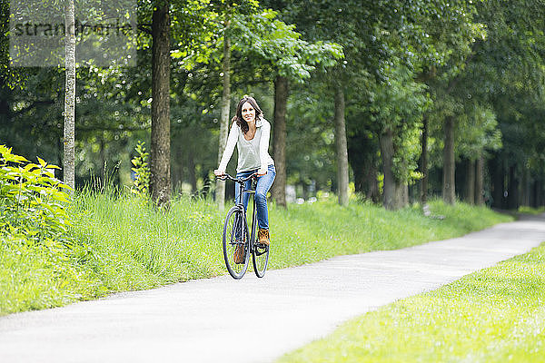 Lächelnde Frau auf dem Fahrrad auf dem Fußweg gegen grüne Bäume im Park