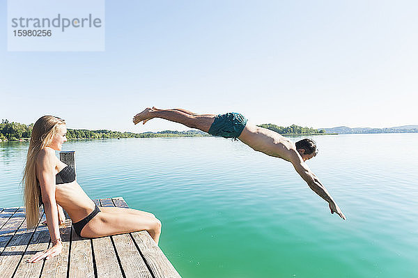 Junger Mann in Badeshorts springt vom Steg in den See  während seine Freundin ihn beobachtet