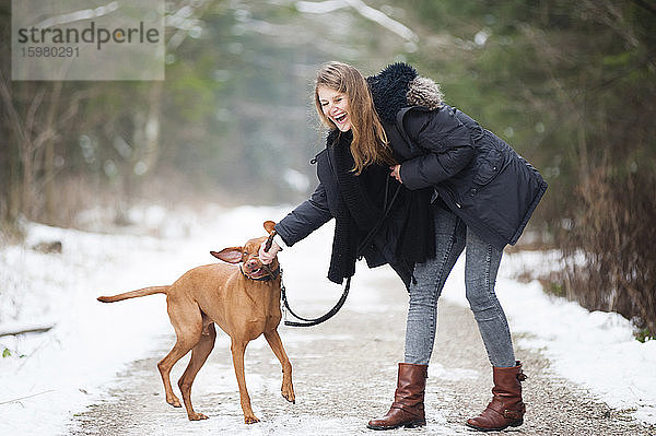 Fröhliche junge Frau spielt mit Hund auf Straße im Wald im Winter