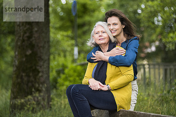 Porträt einer glücklichen Frau  die ihre Mutter umarmt  die auf der Rückenlehne einer Parkbank sitzt