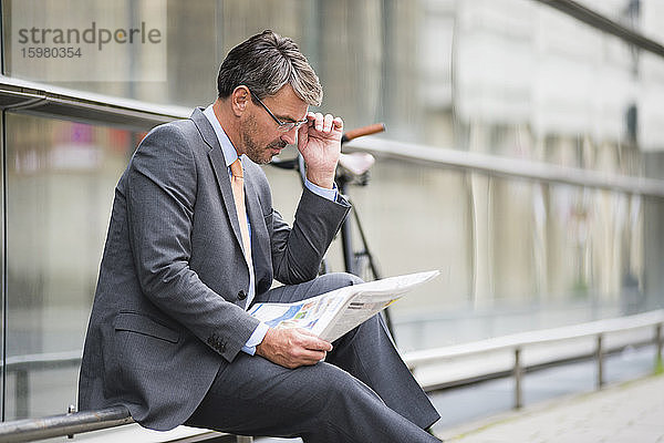 Geschäftsmann mit Brille liest Zeitung und sitzt auf einem Geländer