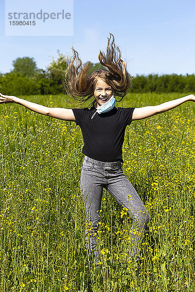 Fröhliches Mädchen mit ausgestreckten Armen und Maske tanzt inmitten von Pflanzen gegen den Himmel