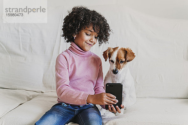 Porträt eines lächelnden kleinen Mädchens  das mit ihrem Hund auf der Couch sitzt und ein Selfie mit dem Smartphone macht