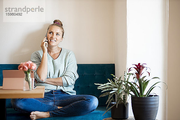 Porträt einer lächelnden blonden Frau am Telefon  die barfuß auf einer Bank zu Hause sitzt