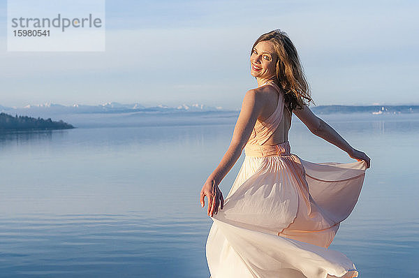 Rückansicht Porträt einer lächelnden schönen Frau im Kleid am Seeufer an einem sonnigen Tag stehend