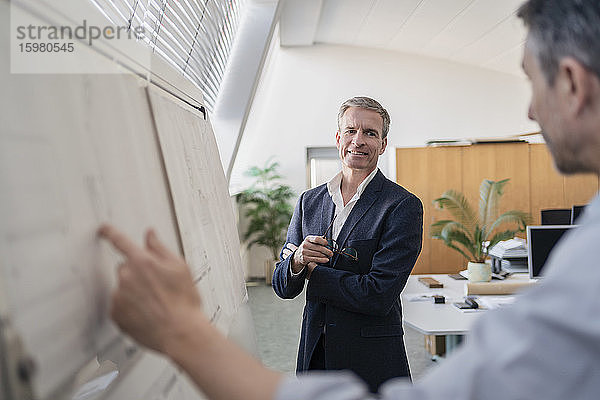 Selbstbewusster reifer Geschäftsmann  der einem männlichen Kollegen zuhört  der während einer Besprechung im Büro eine Strategie am Whiteboard erläutert