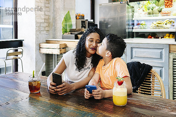Junge küsst Mutter  während er mit Handy und Getränken im Restaurant sitzt