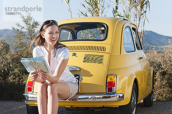 Porträt einer lachenden Frau mit Landkarte  die auf der Stoßstange eines gelben Weinautos sitzt  Sardinien  Italien
