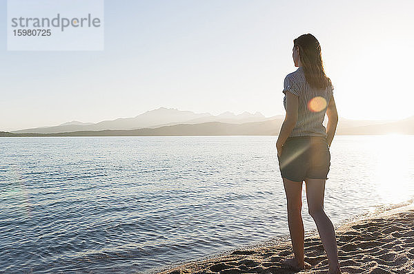 Rückenansicht einer Frau  die bei Sonnenuntergang am Meer steht und die Aussicht betrachtet  Sardinien  Italien