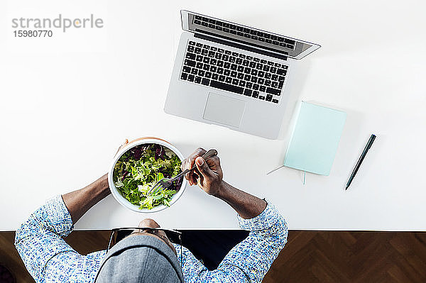 Junger Mann isst Salat  während er mit einem Laptop am Schreibtisch im Büro sitzt