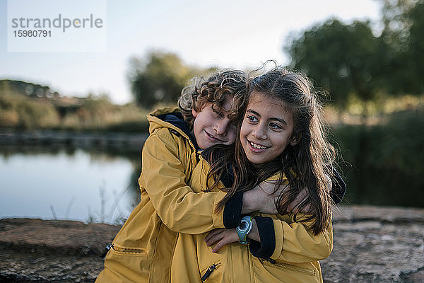 Junge umarmt lächelnde Schwester in gelben Regenmänteln am Flussufer