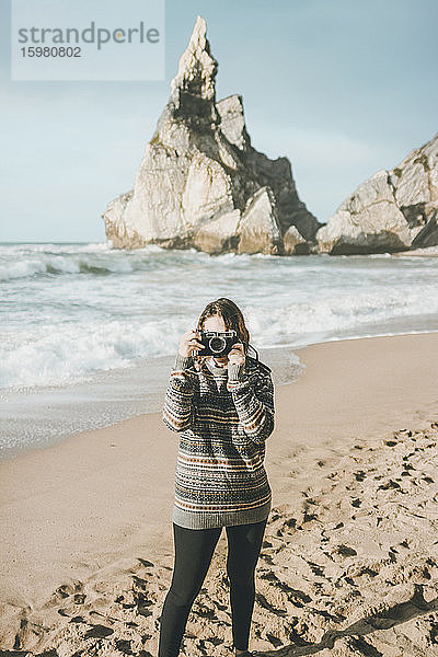 Junge Frau beim Fotografieren  während sie an einer Felsformation am Strand steht  Praia da Ursa  Lisboa  Portugal