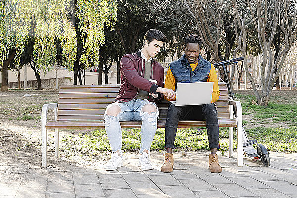 Zwei junge Männer sitzen auf einer Parkbank und teilen sich einen Laptop