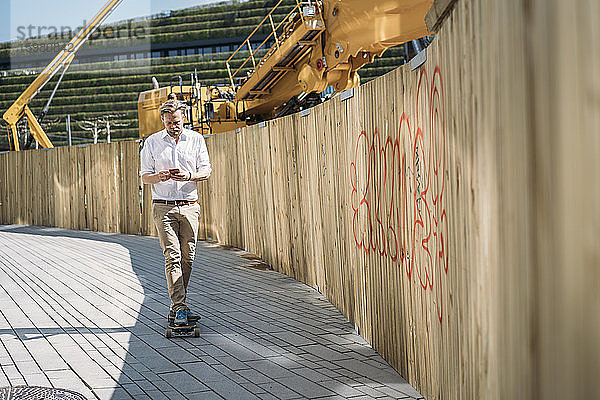Geschäftsmann fährt Skateboard und benutzt sein Smartphone auf einer Baustelle in der Stadt