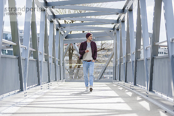 Junger Mann  der mit Ohrstöpseln und Smartphone auf einer Brücke spazieren geht