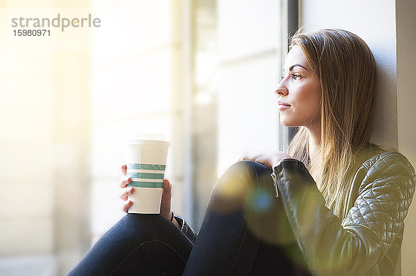 Frau schaut durch das Fenster und hält einen Einweg-Kaffeebecher in einem Café
