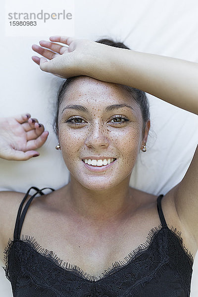 Lächelnde junge Frau mit Sommersprossen im Gesicht liegt auf dem Bett zu Hause