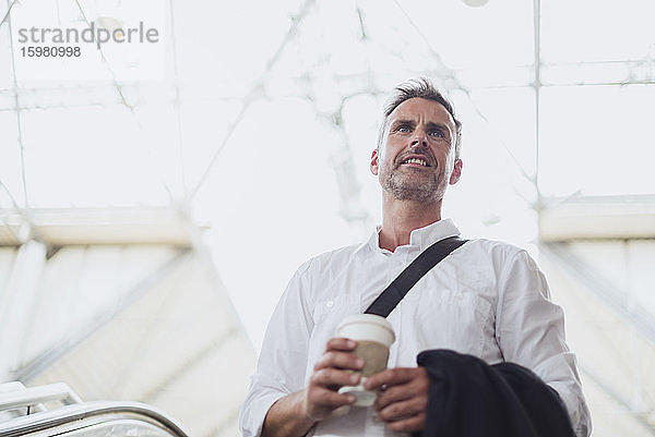 Nachdenklicher Geschäftsmann mit Kaffee in der Hand  der wegschaut  während er auf einer Rolltreppe steht